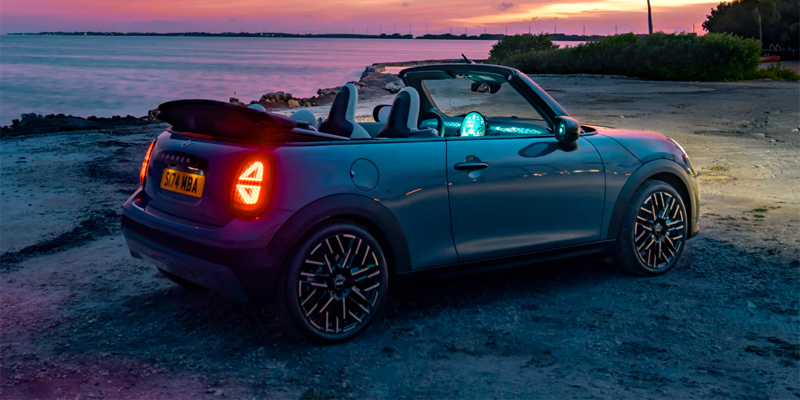 A blue 2025 MINI Cooper convertible car parked by the water at sunset, with reflections and glowing lights.