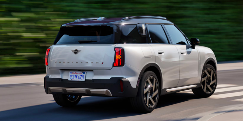 A white 2025 MINI Countryman JCW ALL4 driving on a road with green trees in the background.