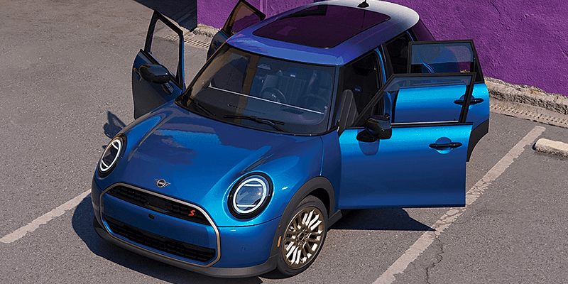A blue 2025 MINI Cooper S 4-Door parked with all doors open, parked on asphalt near a purple wall.