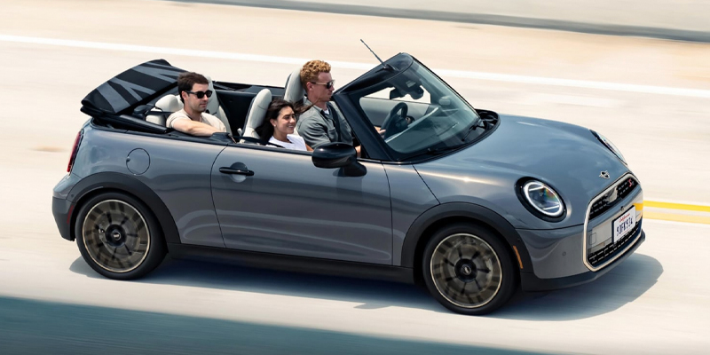 A gray 2026 MINI Convertible Cooper S cruises down the highway with the top down, carrying three passengers enjoying an open-air drive. Available in Morristown, NJ.