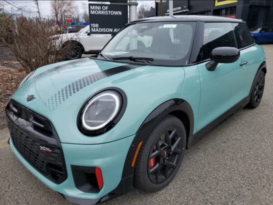 A teal 2026 MINI Hardtop 2-Door JCW parked outdoors, showcasing its sporty design and black accents.