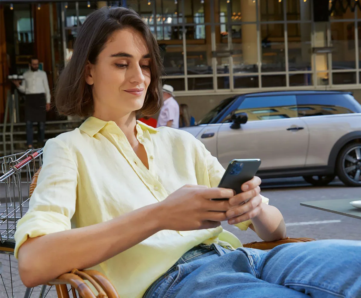 Female wearing yellow collared button-down shirt and jeans, sitting in a chair and holding a smartphone next to a street with a grey MINI vehicle parked in the background. | MINI of Morristown in Morristown NJ
