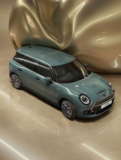 Three-quarter top view of the refined brass front and rear accents on a MINI Clubman Untold Edition, which has a giant CGI, inflating, shiny, brass balloon coming from the side of the car.